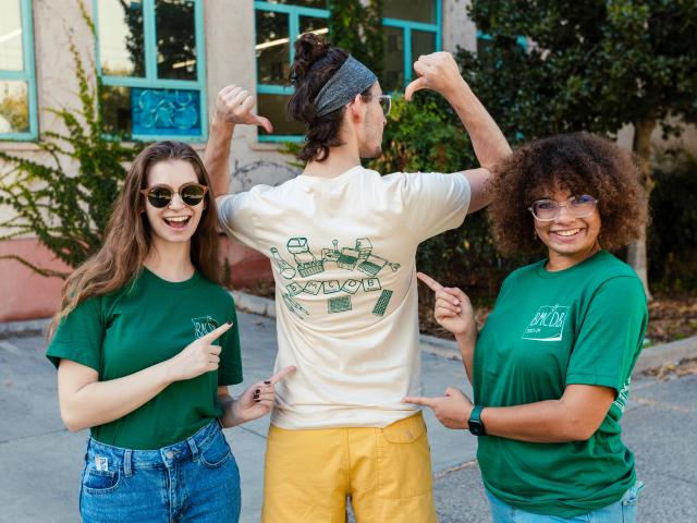 A graduate student stands with his back to the camera, showing off the BMCDB logo on the back of his shirt. Two students stand on either side of him, giving thumbs-up signs and smiling.